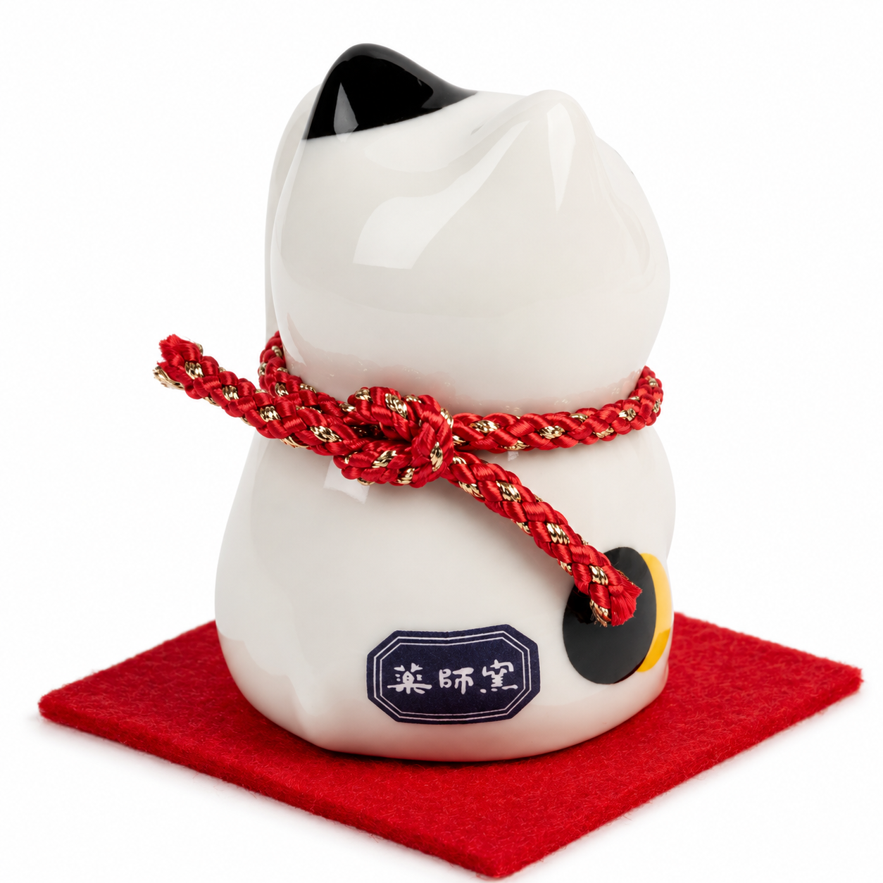 Back view of white calico Maneki Neko lucky cat with red and gold braided collar, Yakushido maker's mark on base, seated on red felt mat