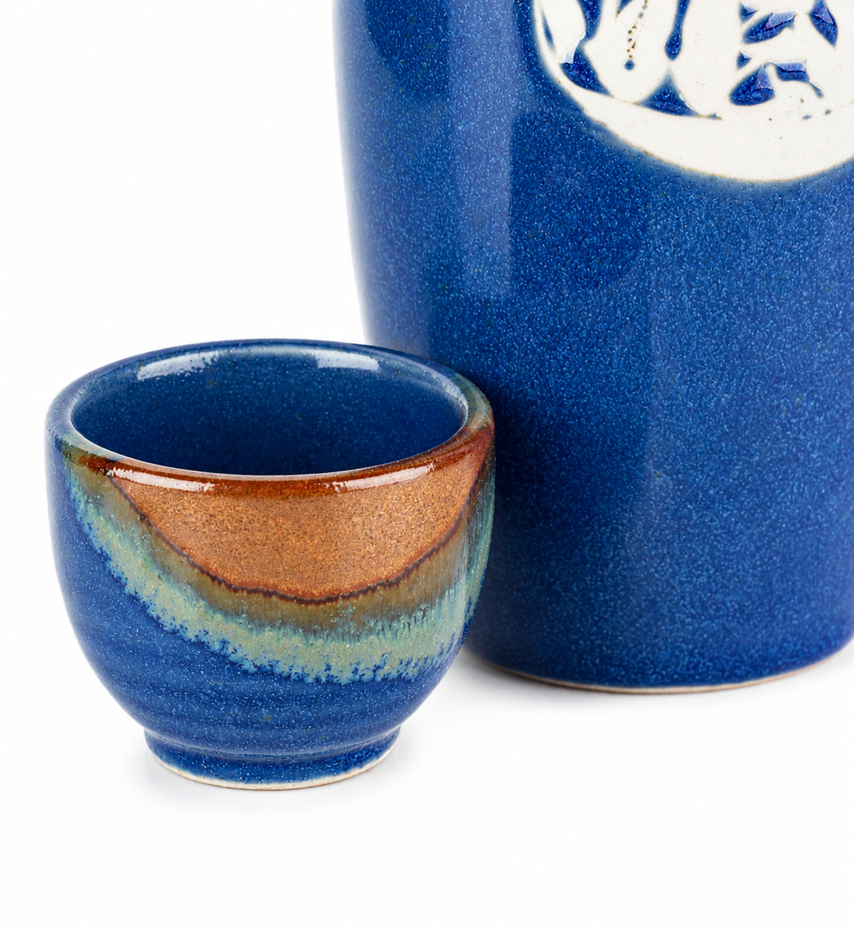 Close-up of Kamakura sake cup showing amber, teal and blue layered glaze on ceramic, beside indigo blue sake jug, made in Japan